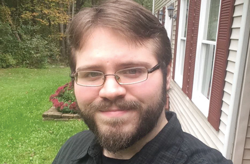 Man smiling wearing glasses and a black collared shirt standing outside of a house.